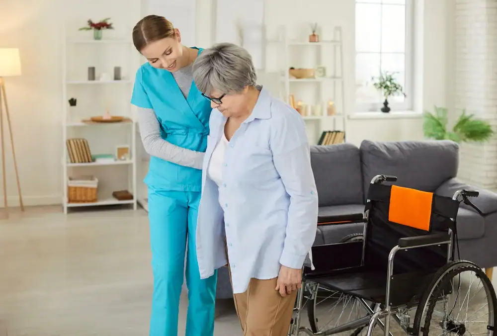 A young caregiver in blue scrubs helps an elderly woman stand up from her wheelchair in a bright NY living room, offering support with a gentle smile—reflecting the care found in occupational therapy Suffolk & Nassau County.