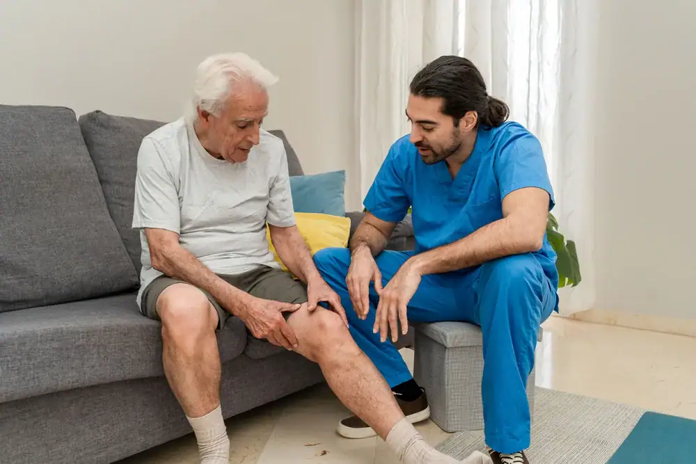 An elderly man seated on a couch touches his knee while talking to a male healthcare professional in blue scrubs—possibly providing physical therapy Suffolk & Nassau County, NY—who kneels beside him and examines the man's knee with concern.