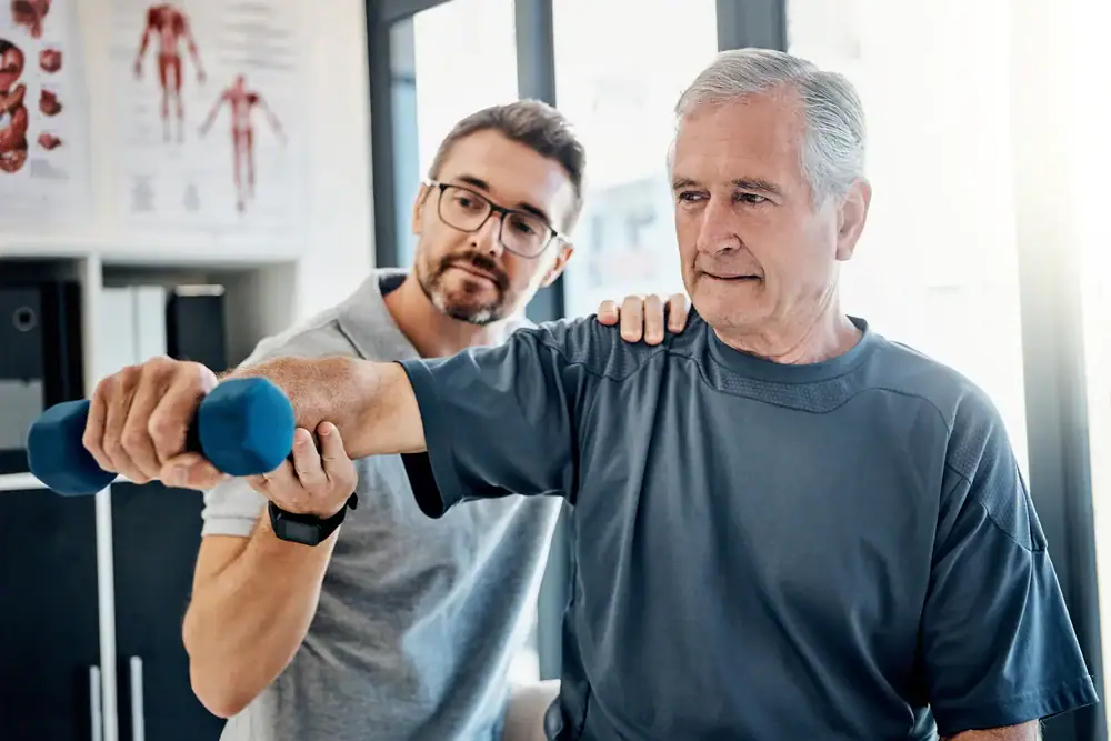 A physical therapist assists an older man lifting a blue dumbbell during a rehabilitation session in a bright, modern clinic. Anatomy posters are visible, highlighting expert physical therapy Suffolk & Nassau County, NY.