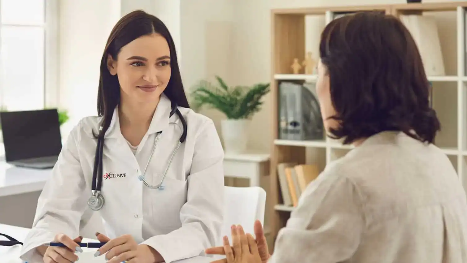 A female doctor in a white coat and stethoscope smiles while discussing occupational therapy Suffolk & Nassau County with a patient across a desk in a bright, welcoming office. Both are engaged in a friendly conversation about treatment options.
