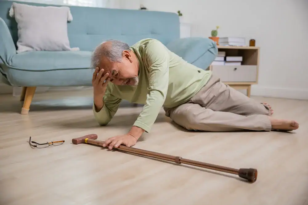 An elderly man is lying on the floor, holding his head in pain, with a cane and eyeglasses beside him. He appears to have fallen in a living room; physical therapy Suffolk & Nassau County may help with recovery and fall prevention.