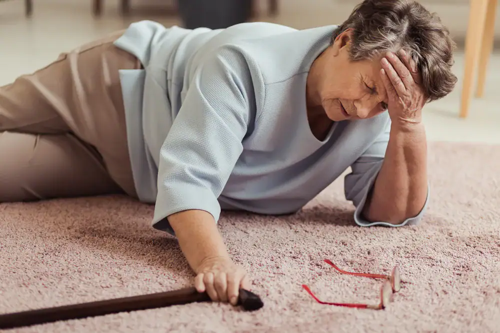 An elderly woman lies on a carpeted floor, looking distressed, with one hand on her forehead and the other reaching for a cane. A pair of glasses lies nearby—she may benefit from physical therapy Suffolk & Nassau County to support her recovery.