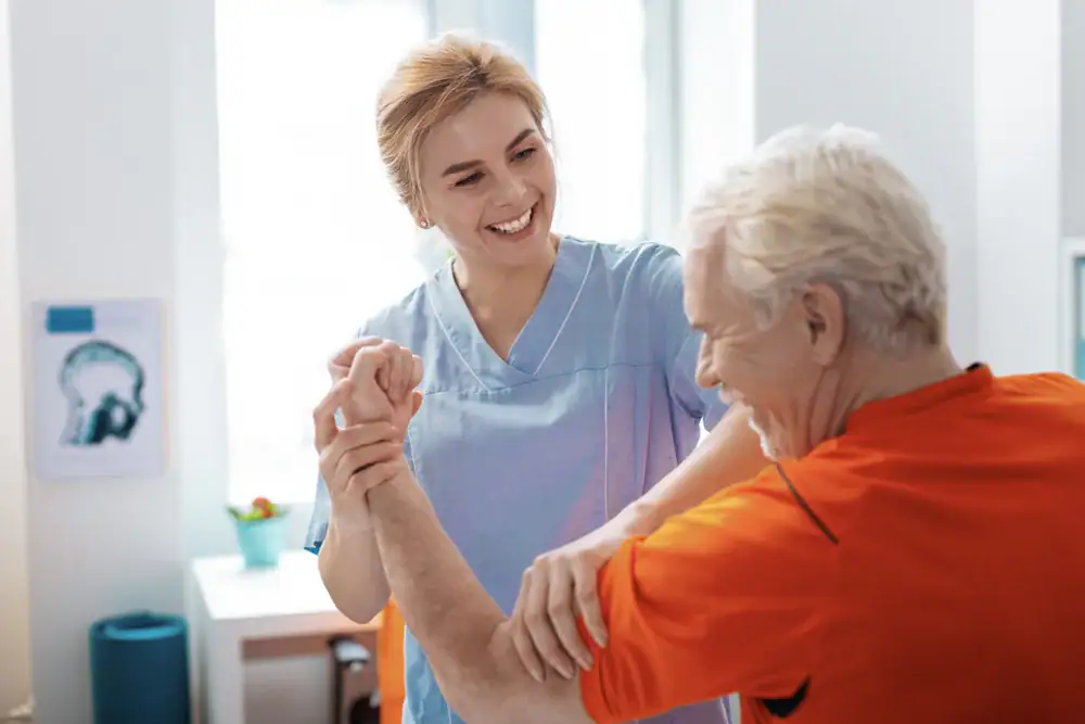 A smiling healthcare professional assists an older man in an orange shirt with arm exercises at a bright NY Physical & Occupational Therapy Suffolk & Nassau County clinic.