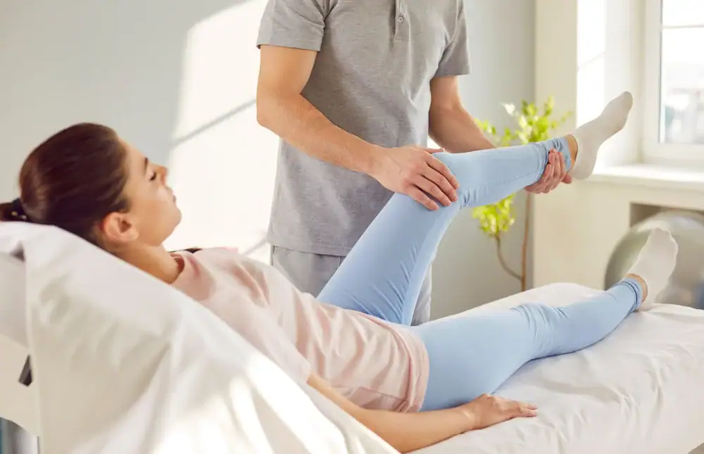 A woman lies on a medical bed while a healthcare professional in a gray shirt helps stretch and examine her bent leg&mdash;likely during a Physical & Occupational Therapy session in Suffolk & Nassau County, NY, in a bright room.