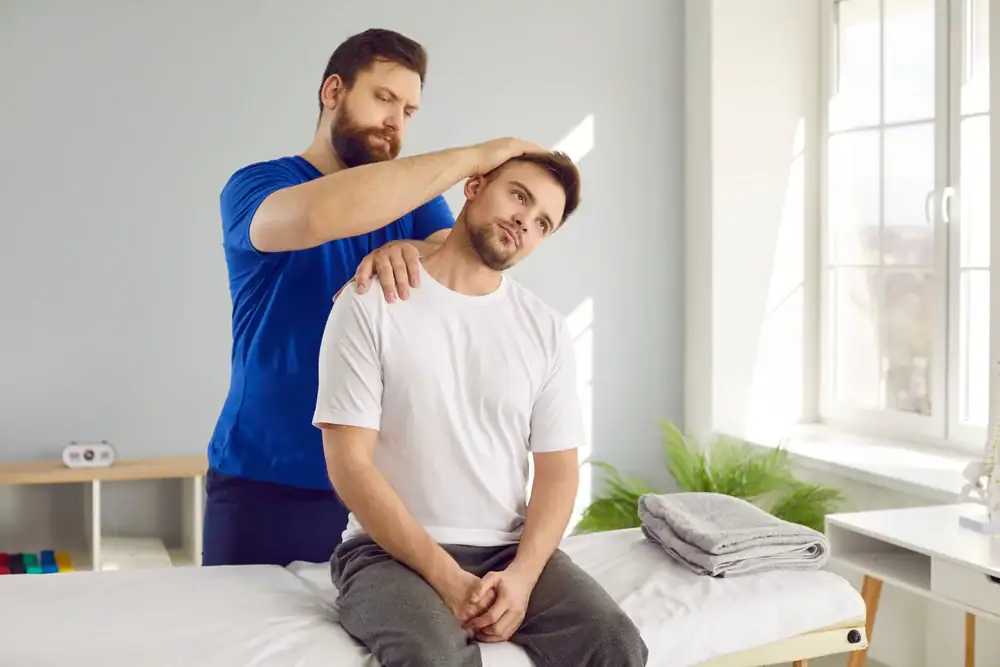 A physical therapist at Physical & Occupational Therapy Suffolk & Nassau County helps a seated man stretch his neck by gently tilting his head to the side in a bright NY therapy room with folded towels and daylight streaming through the window.