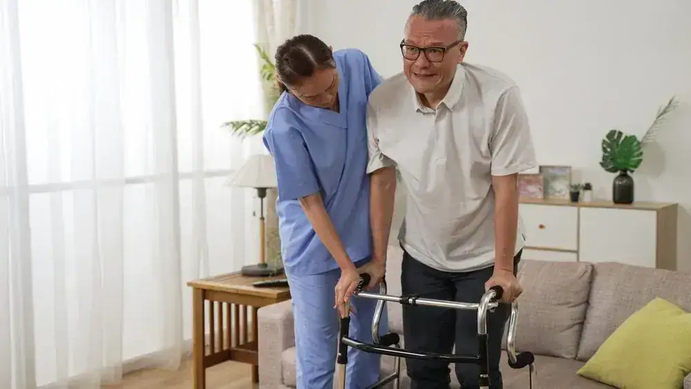 Elderly man using a walker, assisted by a caregiver in a living room.