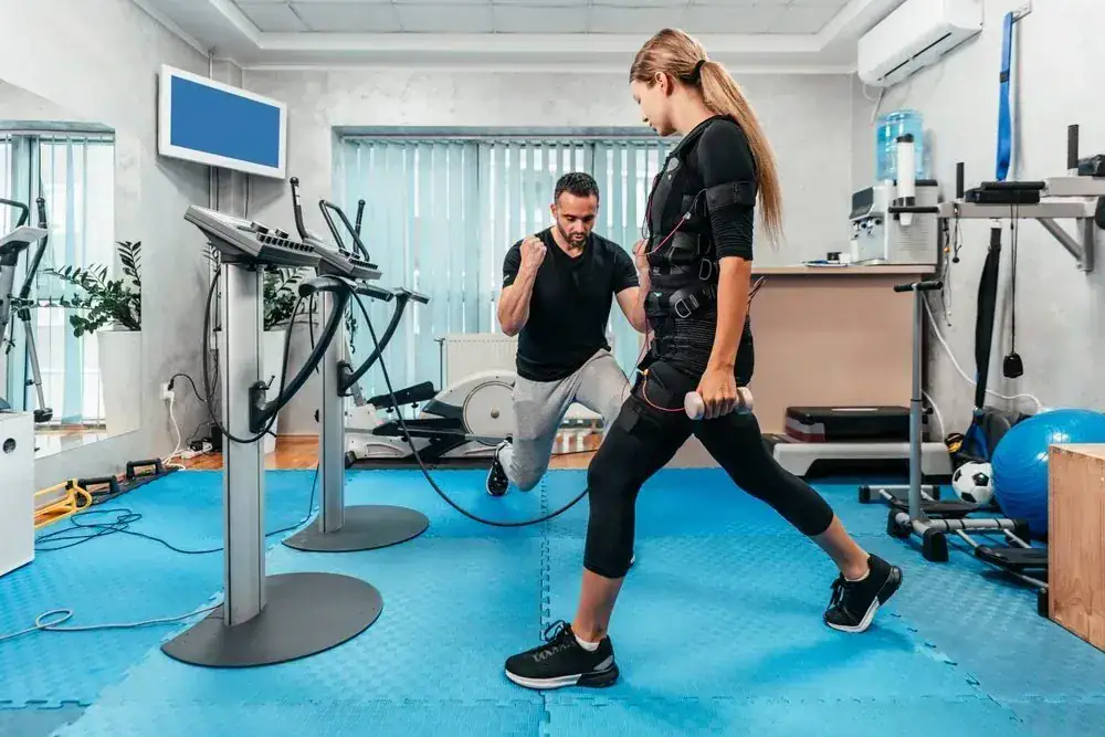 Woman in EMS gear exercising under trainer supervision in a gym.