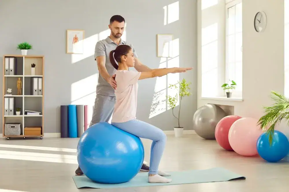 A woman sits on an exercise ball with arms extended forward while a man guides her posture in a bright NY studio, reflecting quality physical therapy Suffolk & Nassau County, surrounded by exercise equipment and large windows.