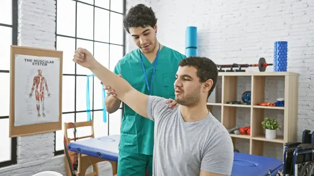 A physiotherapist helps a patient raise his arm during a rehabilitation session in a NY clinic, with exercise equipment and a muscular system chart visible in the background.
