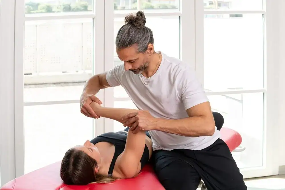 A physical therapist in NY assists a woman lying on a red treatment table by gently stretching her arm, demonstrating expert physical therapy Suffolk & Nassau County care in a bright room with large windows.